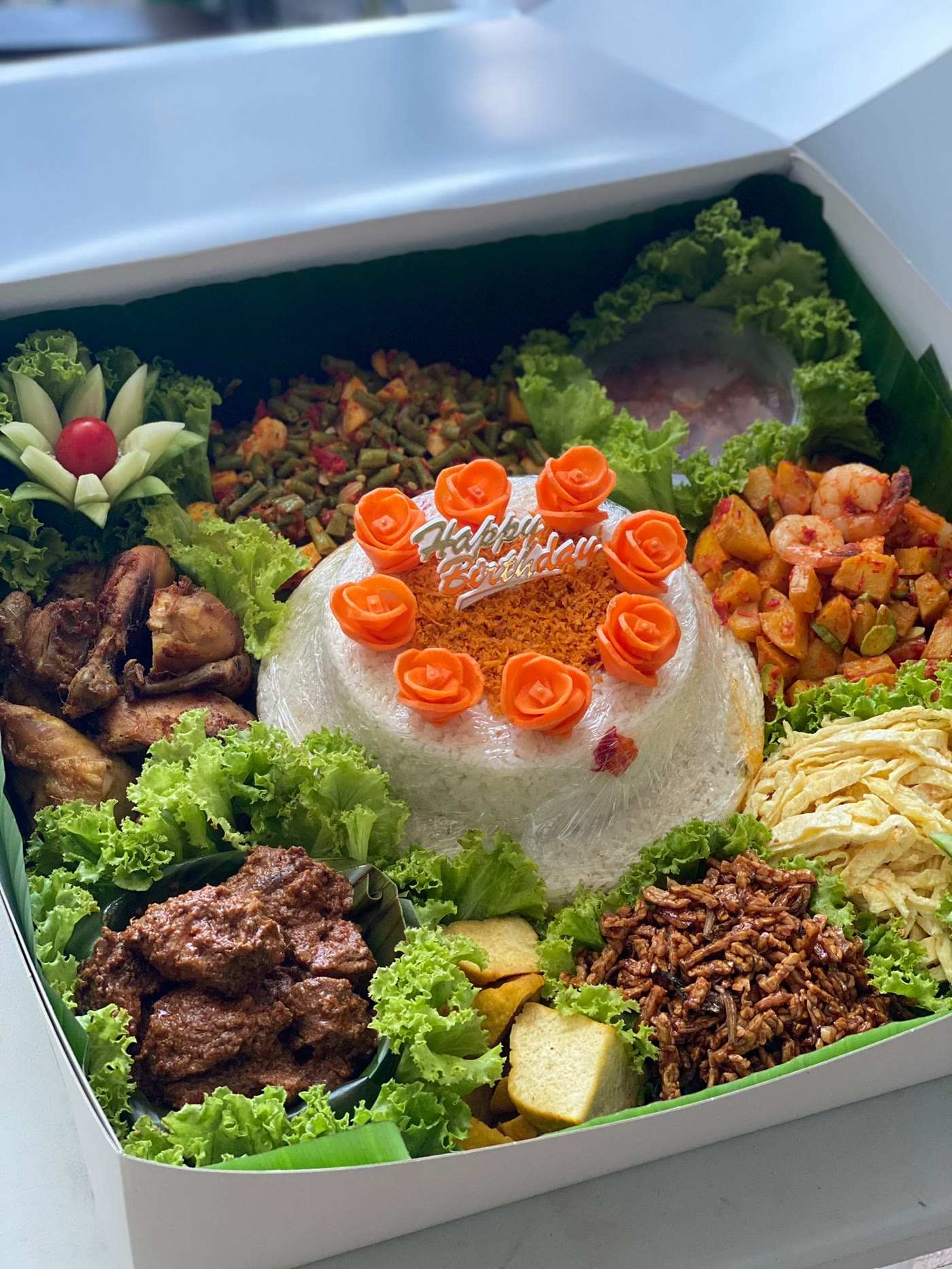 Tumpeng in a gift box with nasi uduk topped with carrot roses, rendang, fried chicken, shredded egg and tempe