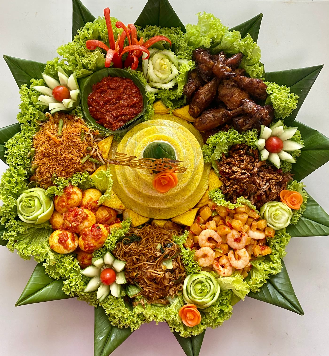 A Riza Kitchen tumpeng on a round tray: a cone of yellow rice in the centre, ringed by chicken, sambal, urap, tempe with bilis, potato with prawn, fried noodles and egg balado, on lettuce and banana leaves